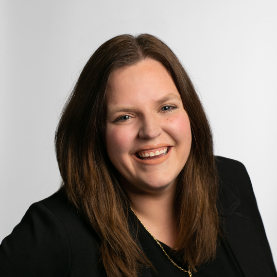 A woman with long brown hair and a black blazer smiles at the camera against a plain white background.