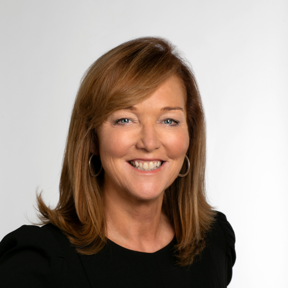 A woman with shoulder-length brown hair, smiling, wearing a black top, and standing against a plain white background.