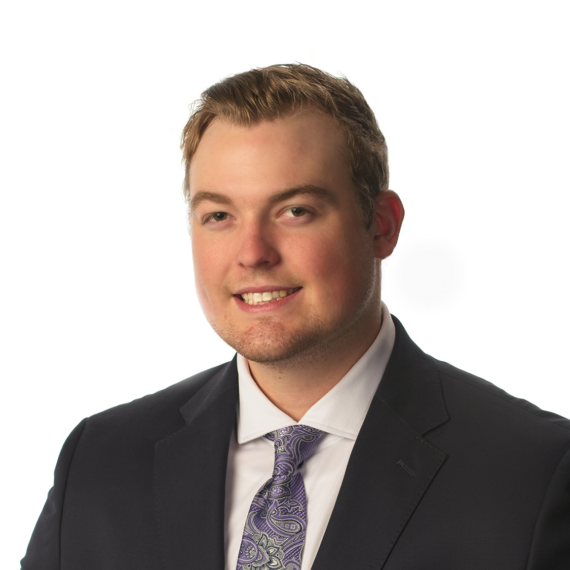 A man in a dark suit, white shirt, and patterned purple tie is posing against a white background. He is smiling slightly while facing the camera.