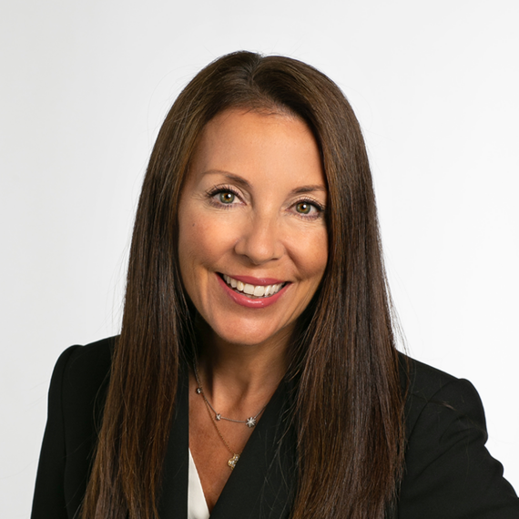 A woman with long brown hair is smiling at the camera, wearing a black blazer and a necklace, standing against a plain white background.