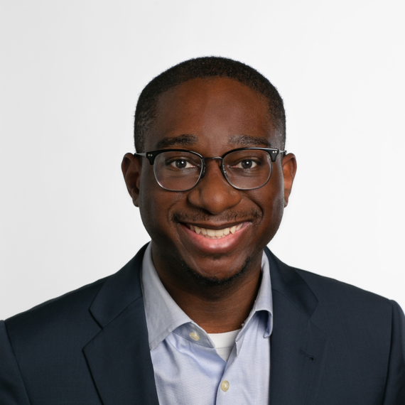 A person with short hair and glasses, wearing a blue blazer over a light blue shirt, smiles at the camera against a plain white background.