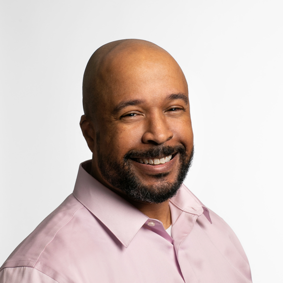 A man with a shaved head and beard, wearing a light pink collared shirt, smiles at the camera against a plain white background.