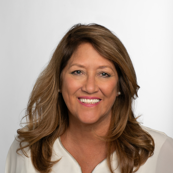 A woman with shoulder-length brown hair and a white blouse smiles while looking at the camera. The background is plain white.