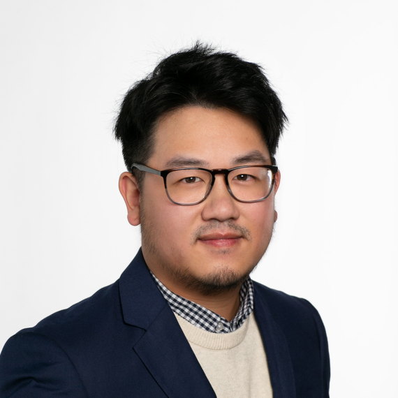 A man with short black hair and glasses, wearing a navy blazer, beige sweater, and checkered shirt, poses against a plain white background.