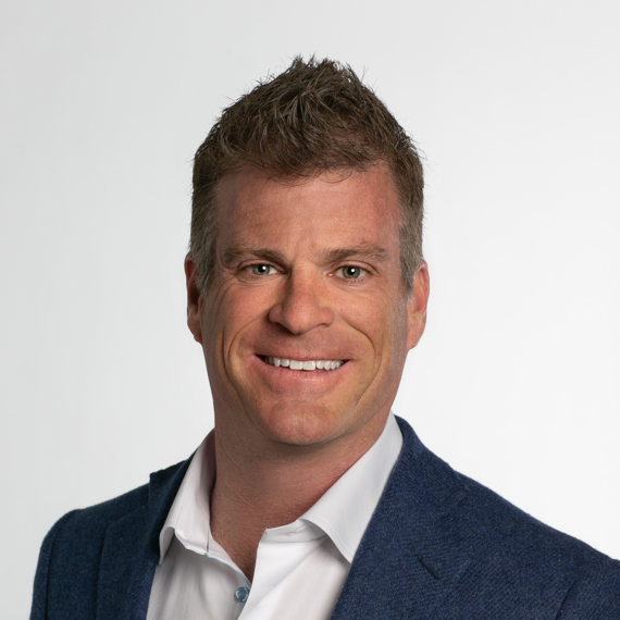 A man with short, spiked hair, wearing a white shirt and a blue blazer smiles at the camera against a white background.