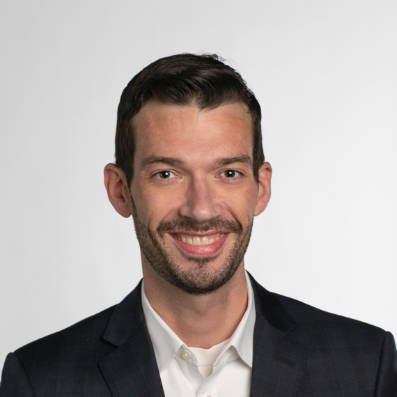 A man with short dark hair, a beard, and a mustache is smiling. He is wearing a dark suit jacket and a white shirt in front of a plain white background.