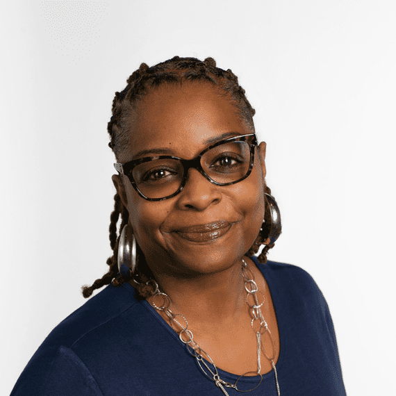 A person with glasses and braided hair, wearing a blue top and a silver necklace, smiles at the camera against a plain white background.