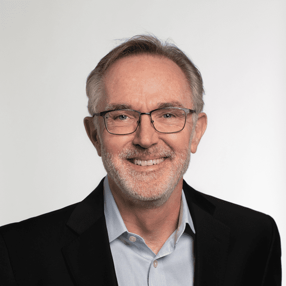 A middle-aged man with glasses, short gray hair, and a beard, dressed in a dark suit jacket over a light blue shirt, smiling against a plain white background.