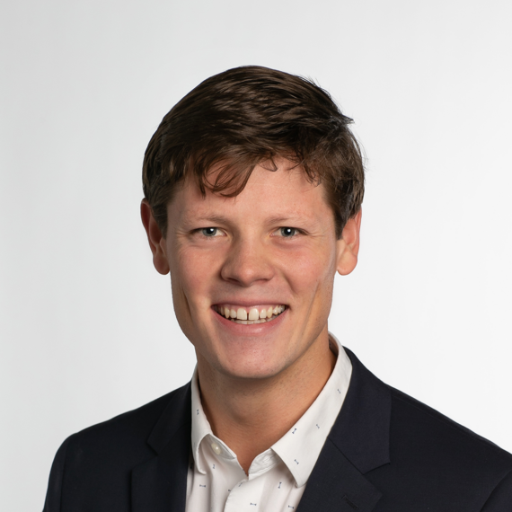 A man with short brown hair, wearing a white shirt and dark blazer, smiles at the camera against a plain white background.