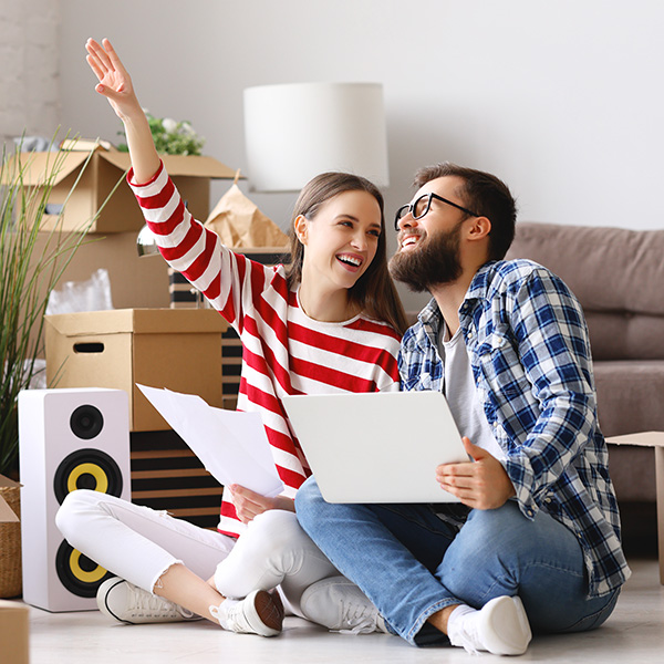 Full body of happy young man with laptop on knees  sitting with wife pointing up while among packed stuff and discussing ideas about interior design of new home