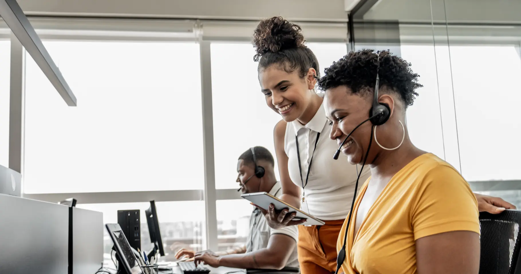 Vrouwelijke-leidinggevende-staat-naast-een-zittende-vrouwelijke-contactcentermedewerker-in-een-callcenter-beiden-glimlachen