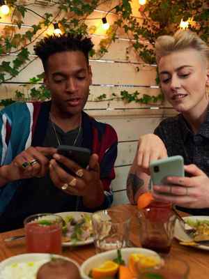 2 happy friends at a restaurant, both are pointing at their phones, showing an example of when you can send money with PayPal.