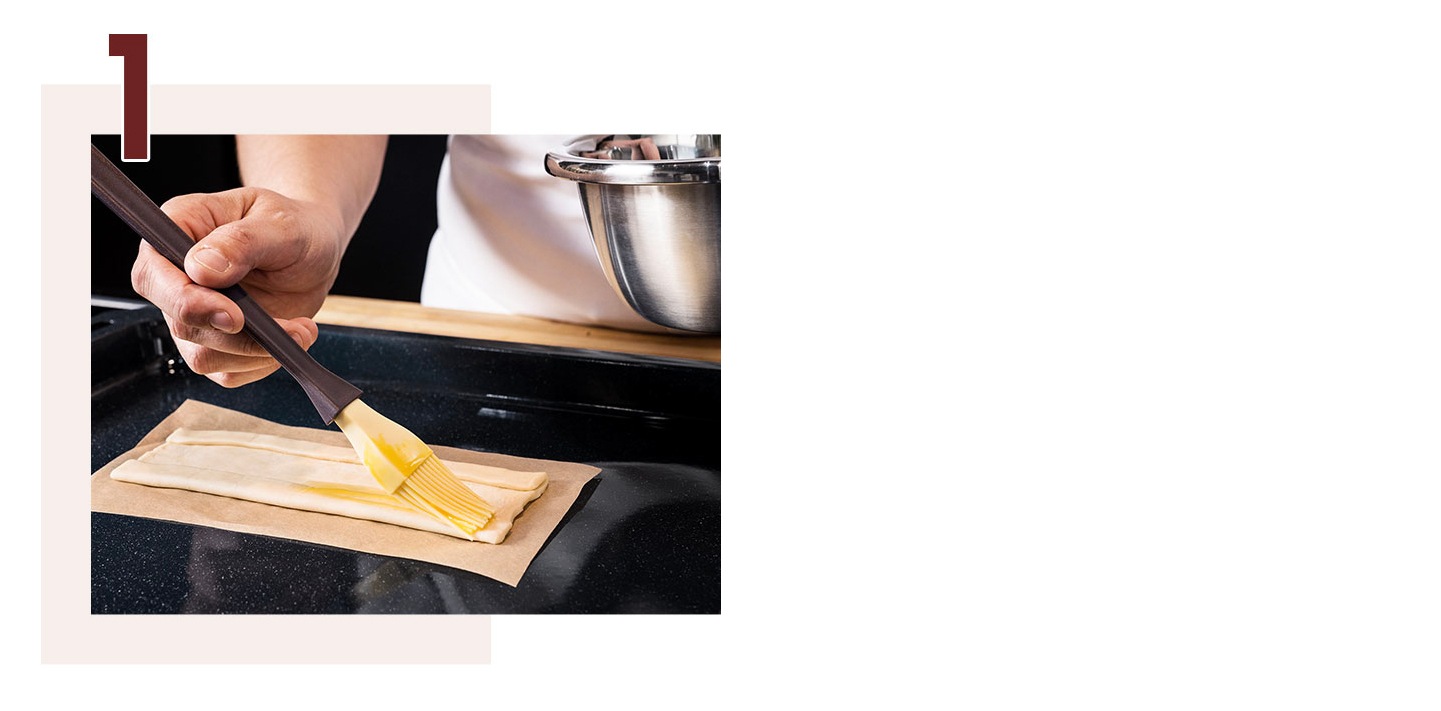 Step 1. A hand is carefully brushing egg wash onto the sides of a pastry using a pastry brush.