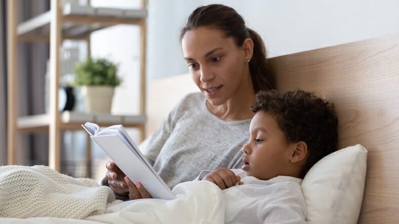 Loving young mom lying in bed with little child reading a story.