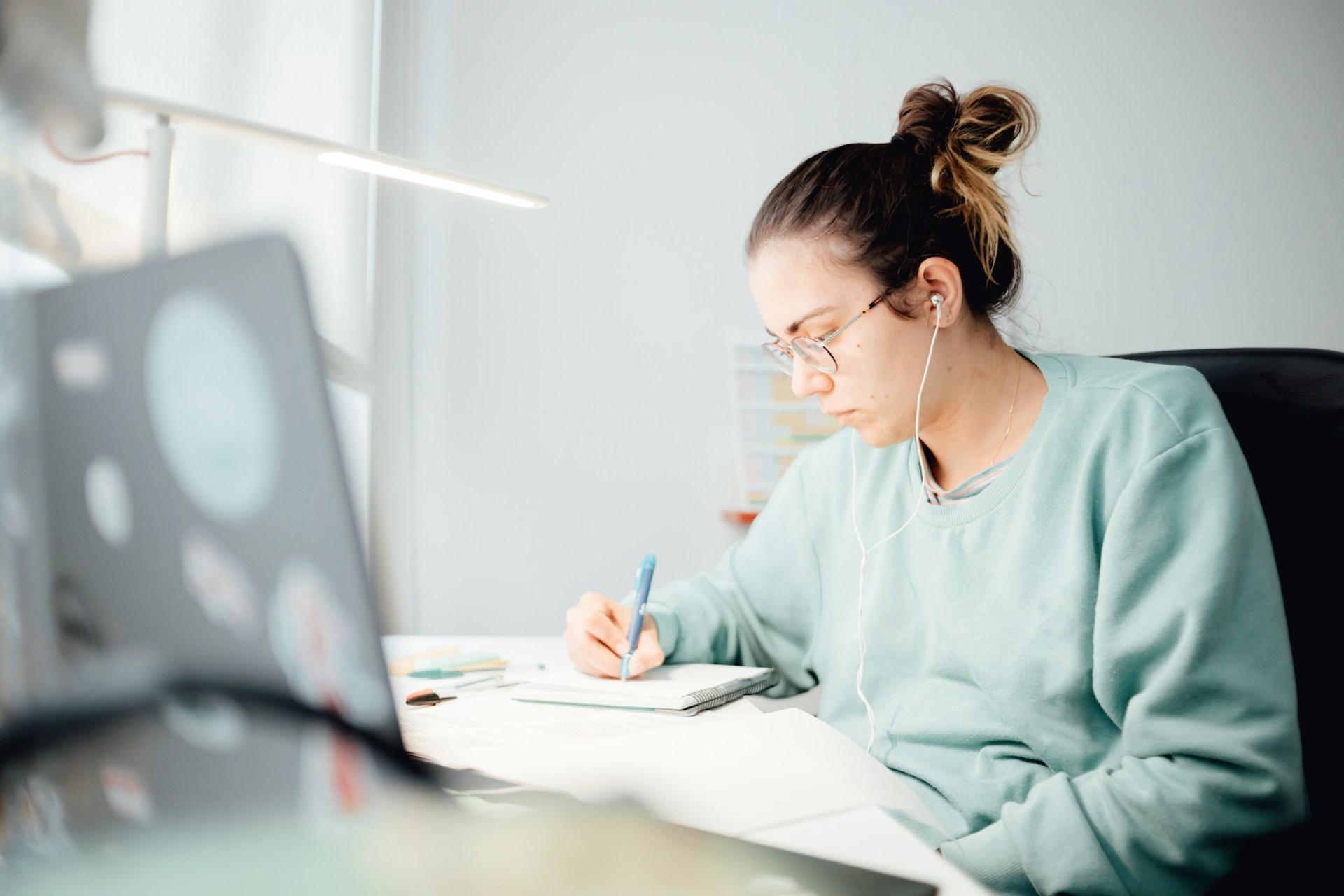 A person wearing a light green shirt and wire framed glasses sits at a table holding a pen in their right hand, writing in the brightly lit space.