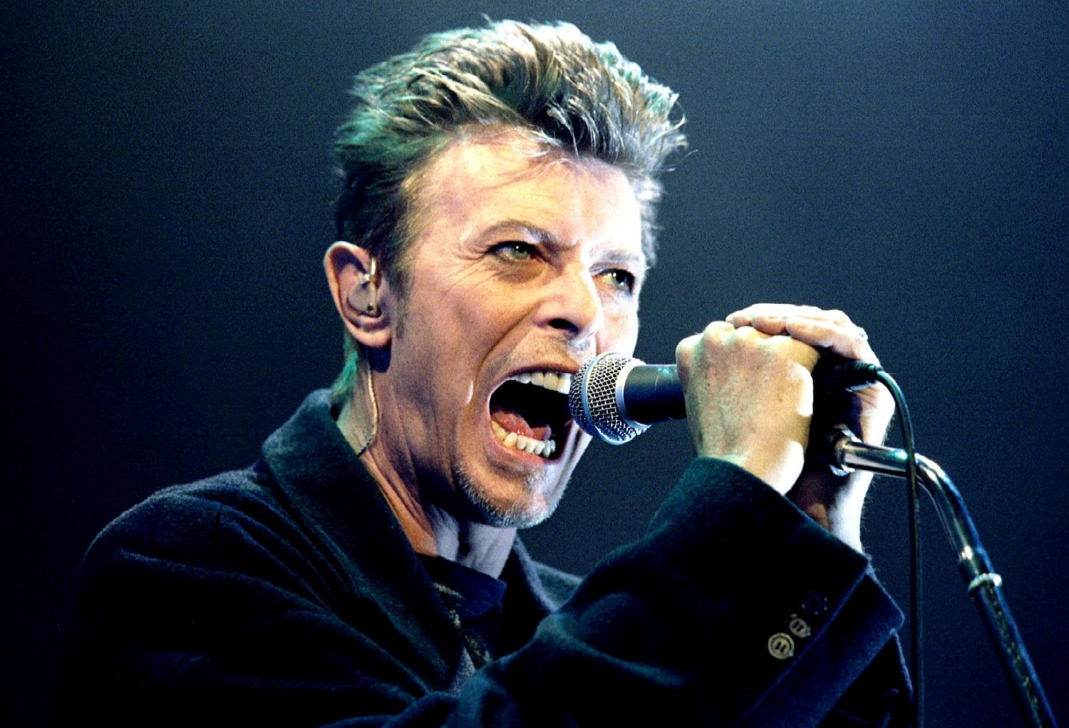 Le chanteur David Bowie en train de chanter les mains sur son micro.