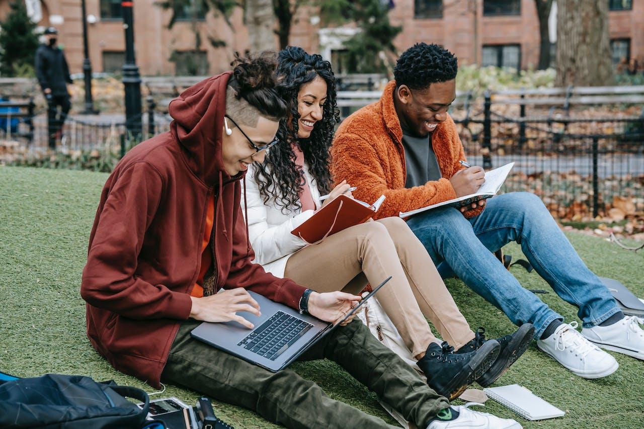 Trois étudiants, deux hommes et une femme sont assis dans l'herbe avec un ordinateur et des carnets pour travailler. 