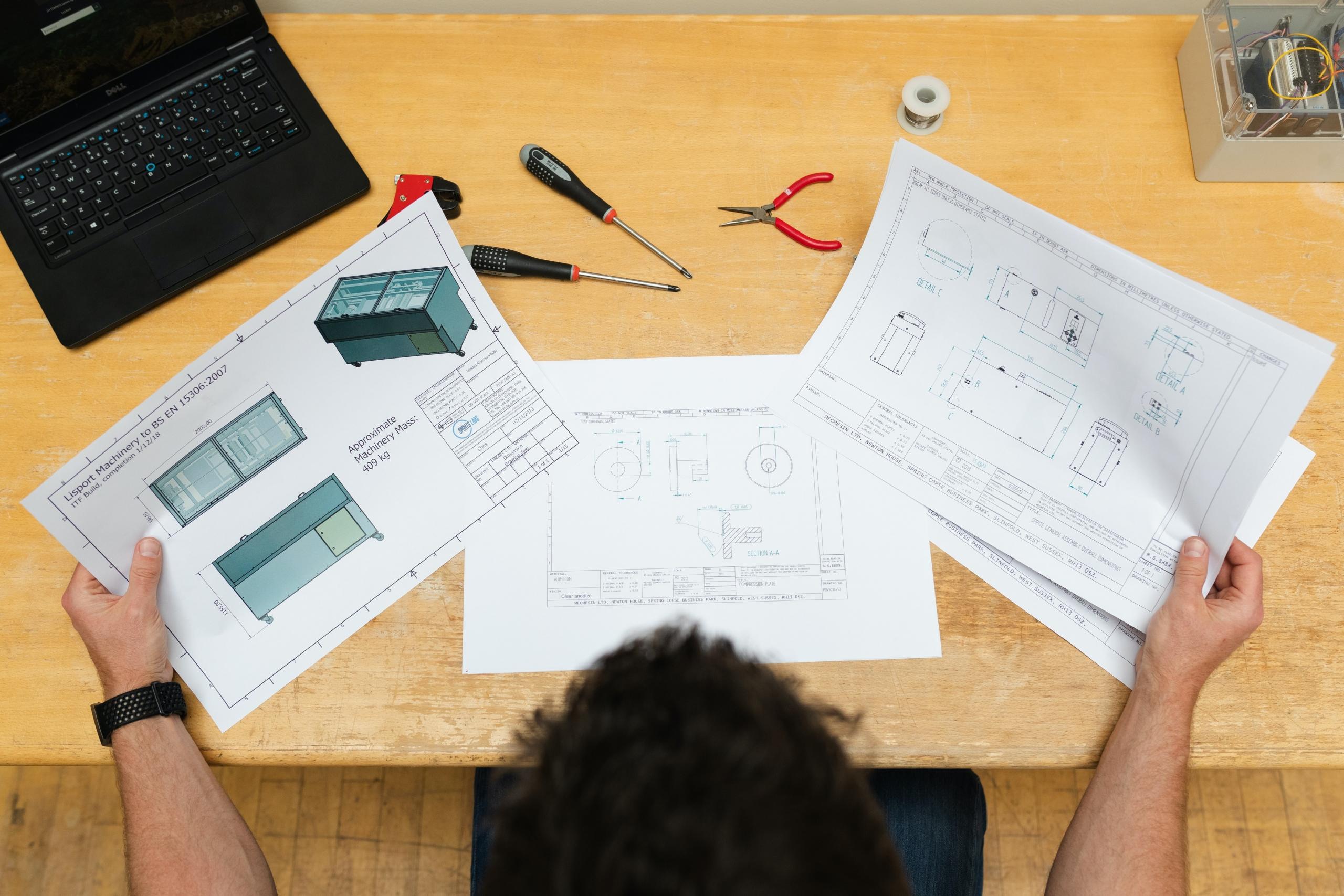 An overhead view of a person sitting at a workbench with various tools scattered in front of them as they scan various papers.