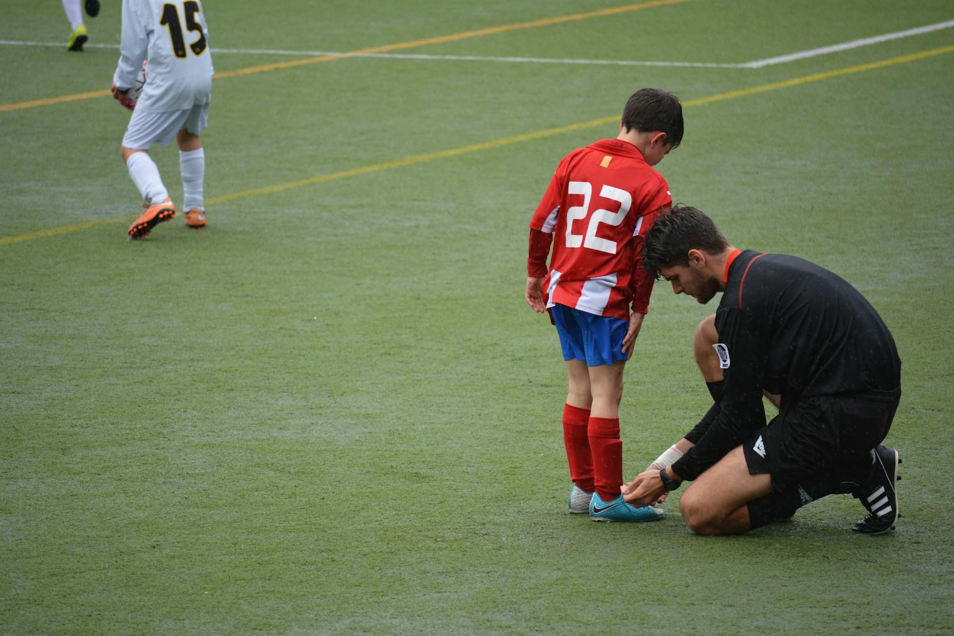 A man wearing black ties a child's shoes on a football pitch.