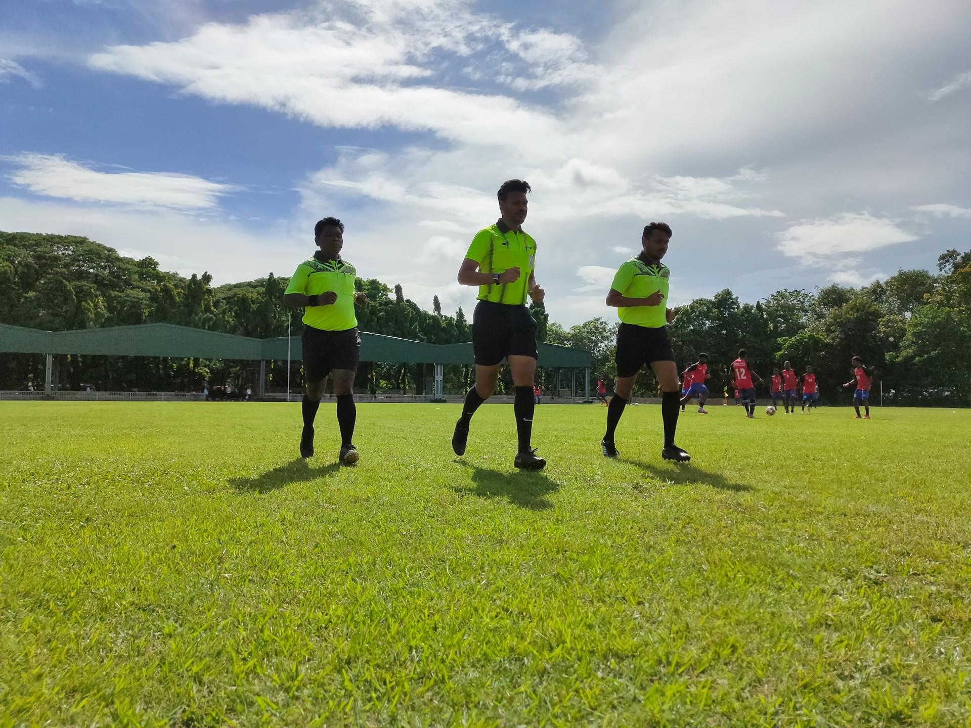 Three men in yellow jerseys run on a football pitch on a partly cloudy day.