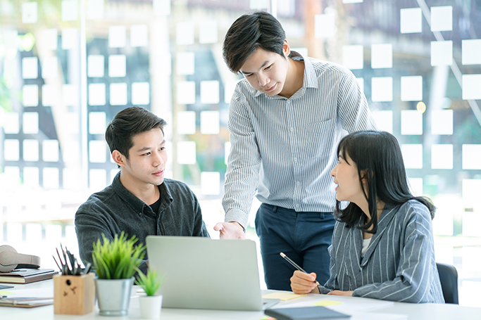 young asian male employee strategizing with two colleagues in front of a laptop