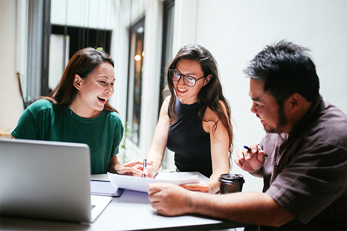 two young female employees with a male colleague laughing together in front of a laptop and paperwork