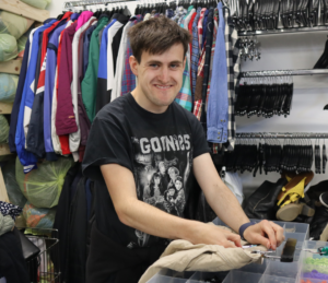 A young man works in Scope organising clothes. He smiles at the camera.