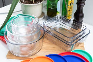 A stack of glass Pyrex bowls, to the left of a stack of rectangular glass Pyrex containers, sitting on a kitchen counter in front of balsamic vinegar, olive oil, and a plant.