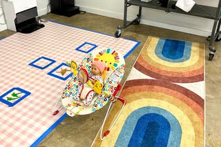 A baby doll resting in a baby bouncer on the floor between a rug and a stained cloth.