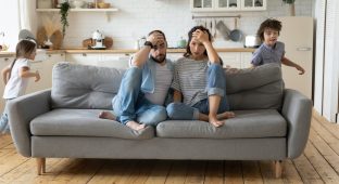 Tired mother and father sitting on couch feels annoyed exhausted while noisy little daughter and son shouting run around sofa where parents resting. Too active hyperactive kids, need repose concept