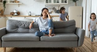 Exhausted young mum sit on couch in kitchen feel unwell tired from ill-behaved loud little children running playing, sick annoyed mother or nanny relax on sofa suffer from headache, parenting concept