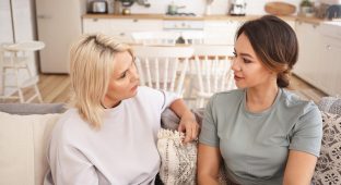 People, lifestyle, family, love and relationships concept. Indoor shot of blonde mature mother sitting on sofa with her young brunette daughter, talking, spending nice time together, drining tea