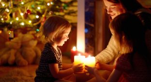 Young mother and her two little daughters sitting by a fireplace holding candles in a cozy dark living room on Christmas eve