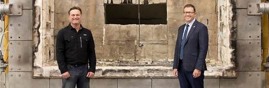 Two white males, one in a black sweater and jeans, and one in a suit and tie, stand smiling in front of a fire test furnace