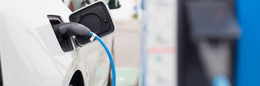 White electric car with charging cord attached; charging station in right background