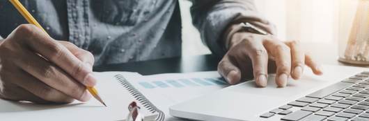 Closeup view of desk while man writes in notebook with other hand on computer with glasses and calculator nearby.  