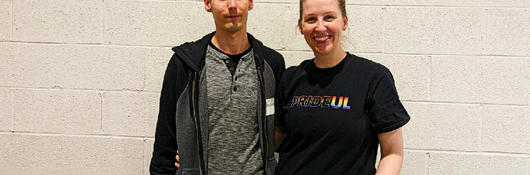 Man and woman from UL PRIDE smile while standing in front of white wall at volunteer event. 