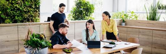 In a modern office with a green plant wall and windows, a group of young co-workers collaborate around a modern wooden dining table.