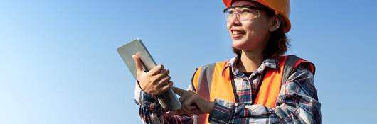 Female engineer in orange safety gear tests solar panel with tablet. 