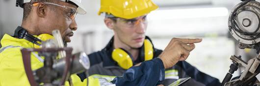 Factory line workers in safety gear examine and discuss a machine part.