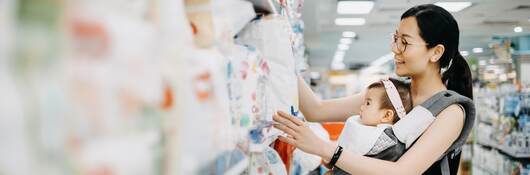 Young Asian mother carrying cute baby girl shopping for baby product in a shopping mall and is looking at a variety of diapers.