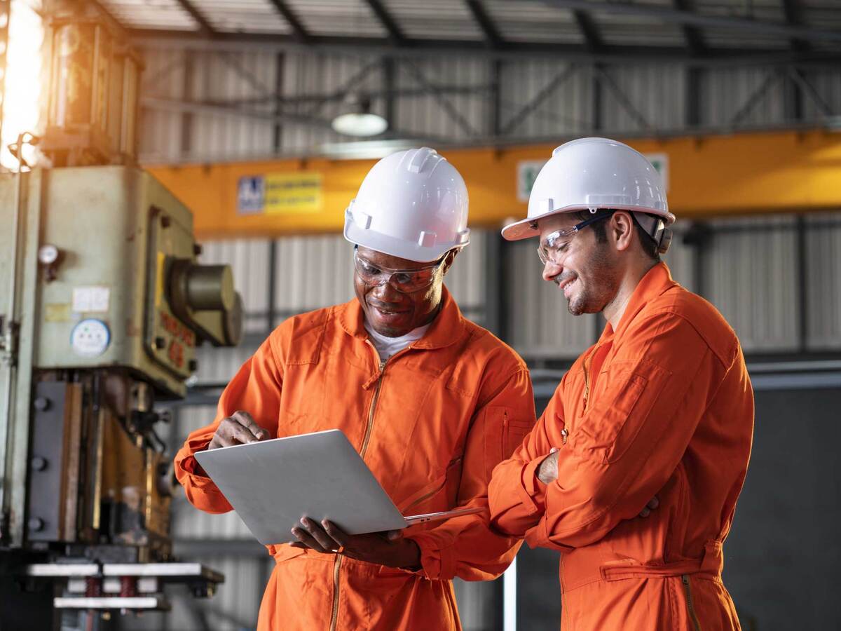 Two engineers in orange coveralls and white hardhats looking at a laptop