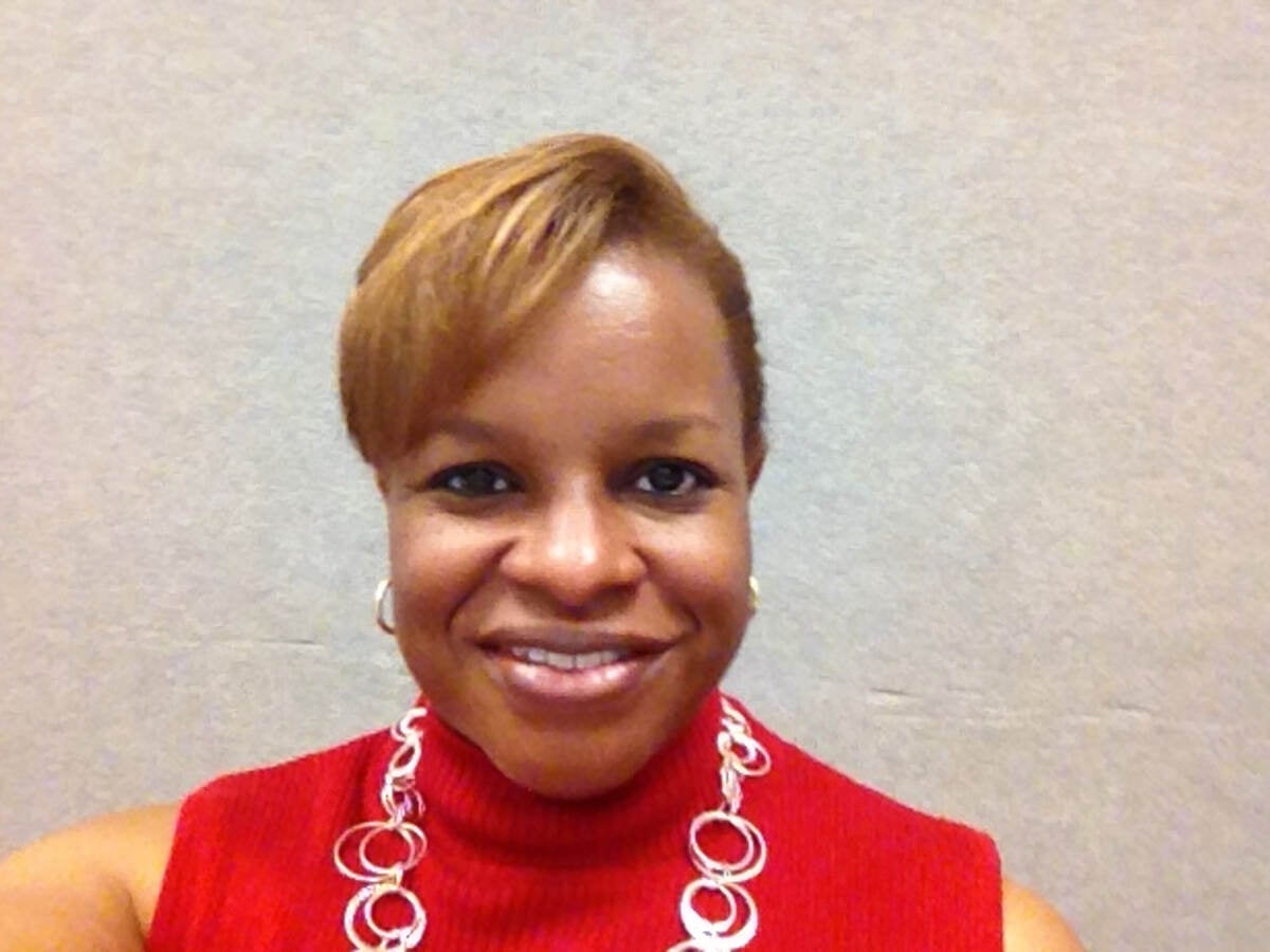 Kim Brooks in red shirt and silver necklace by light brick wall. 