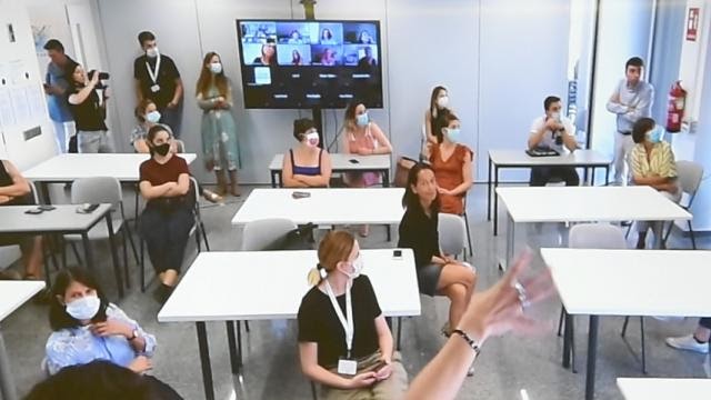 Classroom of college students wearing masks with video conference showing gallery view of remote students at the back of the classroom