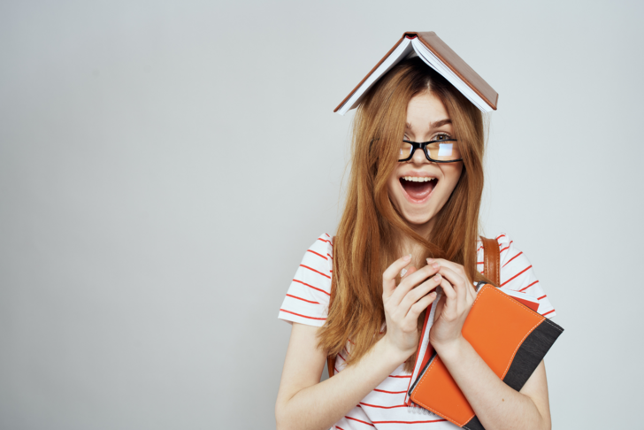 A smiling woman clutches two books in her arms while one book is perched on her head like a hat.