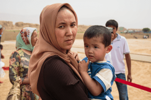 A woman wearing a beige head scarf is pictured looking over her shoulder and holding a toddler in her arms wearing a blue and white stripped t-shirt.