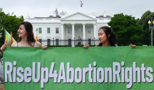 The image shows two women holding a large green banner with the text "RiseUp4AbortionRights" written in bold white letters. They are standing in front of the White House, which is visible in the background. The image appears to be taken during a protest or demonstration advocating for abortion rights. The women are smiling, and the atmosphere suggests an organized and peaceful protest.