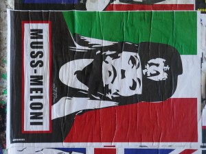 The image depicts a poster with a stylized black-and-white illustration of a woman wearing a military-style uniform and cap, reminiscent of fascist propaganda art. The background features the Italian flag with vertical green, white, and red stripes. Below the image, in bold black and red text, is the label "MUSS-MELONI", which is a play on the names of Benito Mussolini, the Italian fascist dictator, and Giorgia Meloni, the Italian politician. The image appears to be a piece of street art or protest art, criticizing Meloni by drawing a parallel between her and Mussolini.