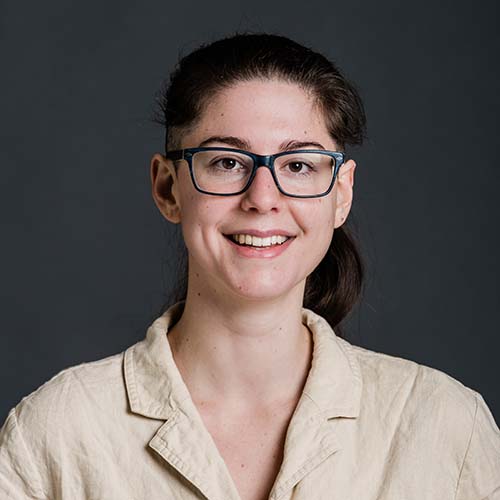 Photo of Dana Ferrante, a young white woman with long brown hair and an undercut. She smiles, wears purple glasses, and a sand-colored shirt.