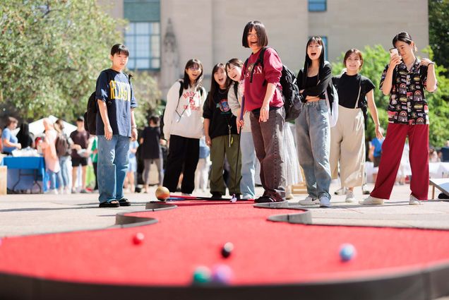 Photo: A group of college students wearing casual clothing stand outside throwing bean bags at a board with a hole in it on a sunny day